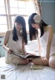 Two young women sitting on a bed reading a book.